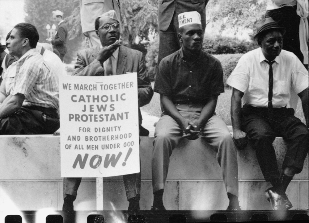 The civil rights march from Selma to Montgomery, Alabama in 1965. Photograph shows some participants in the civil rights march sitting on a wall resting, one holds a placard which reads, "We march together, Catholics, Jews, Protestant, for dignity and brotherhood of all men under God, Now!"